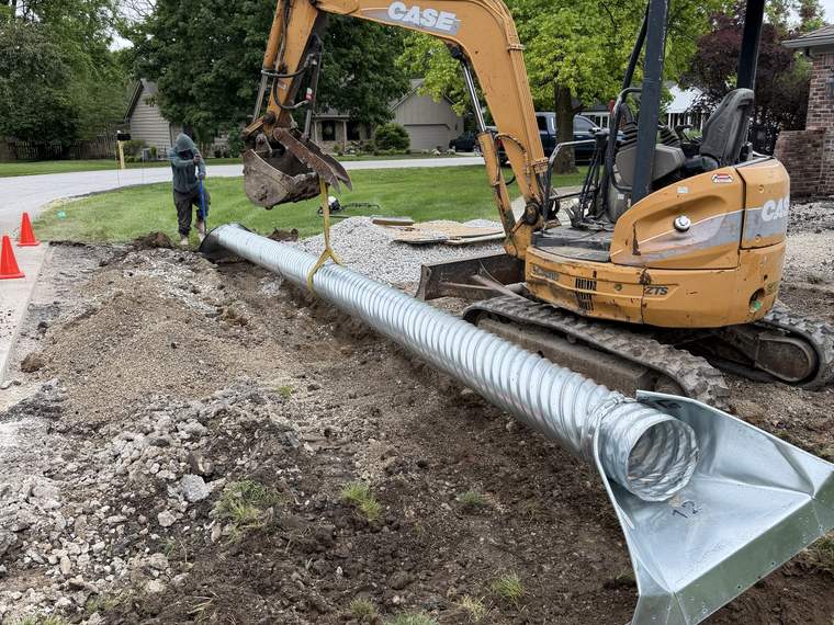 Corrugated metal culvert staged beside an open trench with a compact excavator