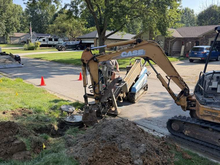 Compact excavator digging a utility trench with locate flags marked across the lawn
